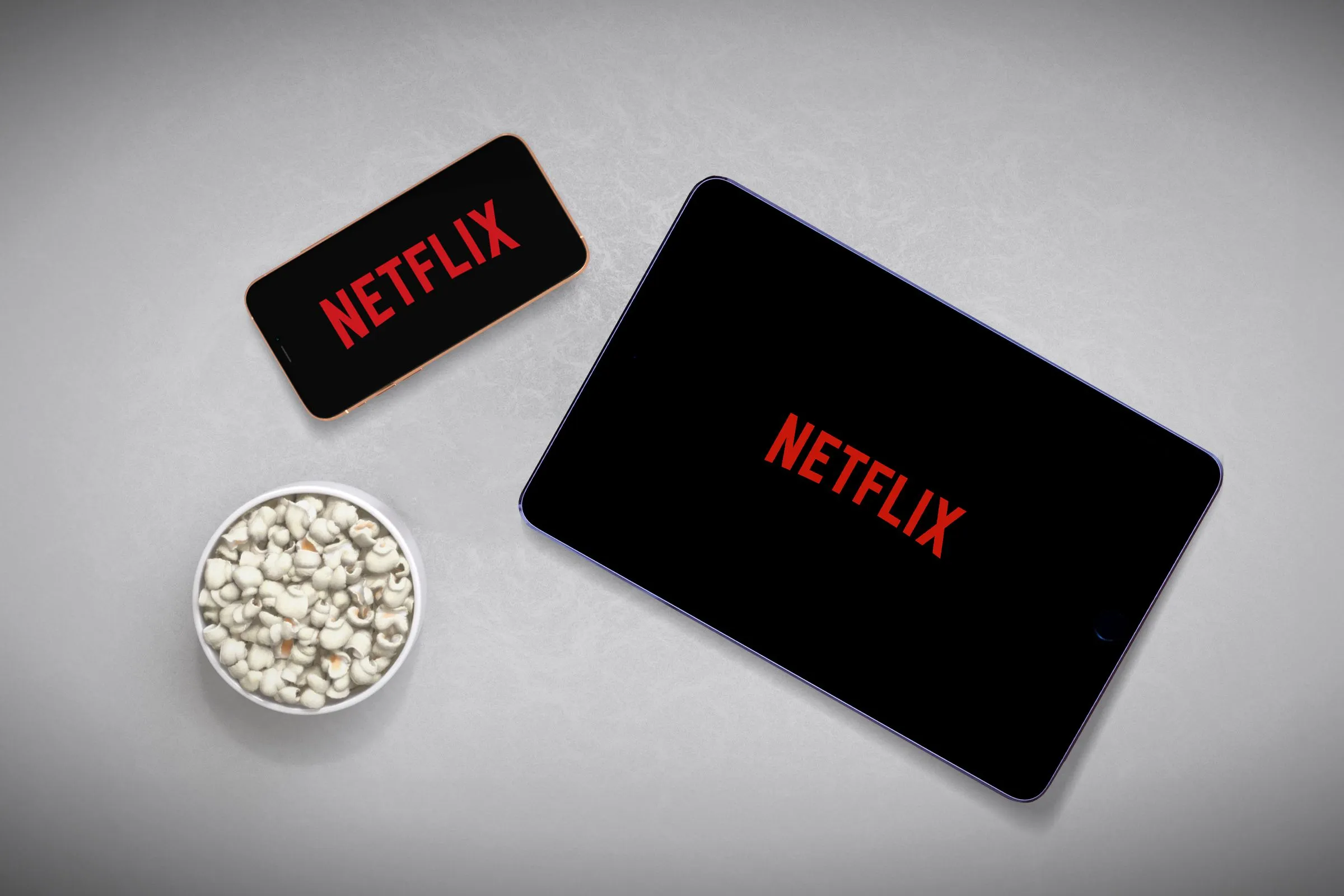 Een smartphone naast een tablet, beide met Netflix, met popcorn in de hoek